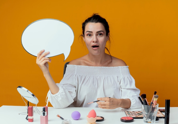 Foto gratuito hermosa niña sorprendida se sienta a la mesa con herramientas de maquillaje tiene burbuja de chat y pincel de maquillaje aislado en la pared naranja
