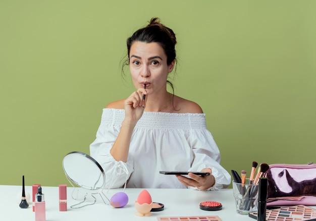 Hermosa niña sorprendida se sienta a la mesa con herramientas de maquillaje tiene paleta de sombra de ojos y pincel de maquillaje aislado en la pared verde