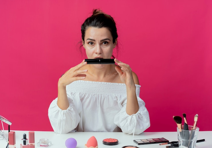 Foto gratuito hermosa niña sorprendida se sienta a la mesa con herramientas de maquillaje tiene rímel aislado en la pared rosa