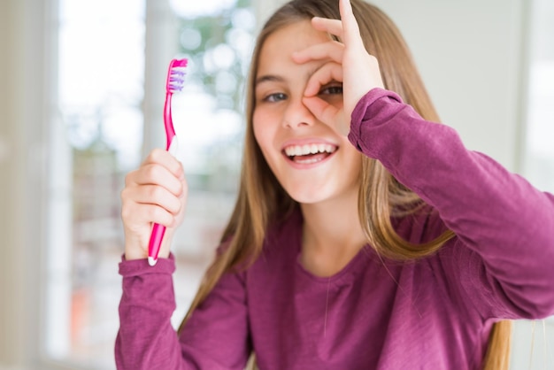 Foto gratuita hermosa niña sosteniendo un cepillo de dientes dental rosa con cara feliz sonriendo haciendo el signo de ok con la mano mirando a través de los dedos