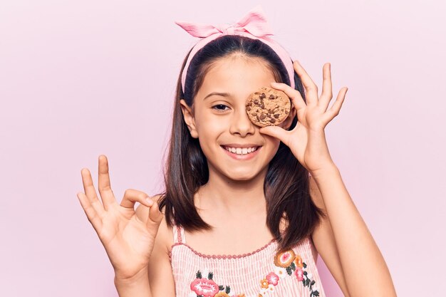 Hermosa niña sosteniendo galleta haciendo el signo de ok con los dedos sonriendo amigable gesticulando excelente símbolo