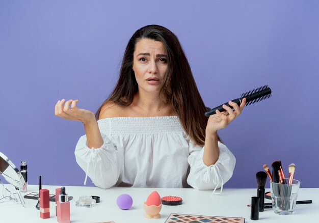 Hermosa niña triste se sienta a la mesa con herramientas de maquillaje con peine aislado en la pared púrpura