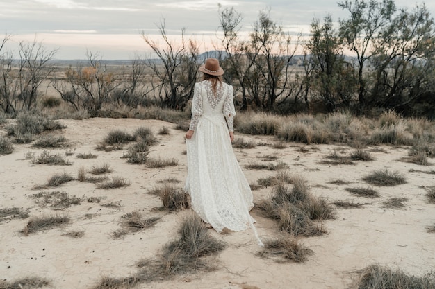 Hermosa novia caminando en una tierra con arbustos y árboles secos al atardecer