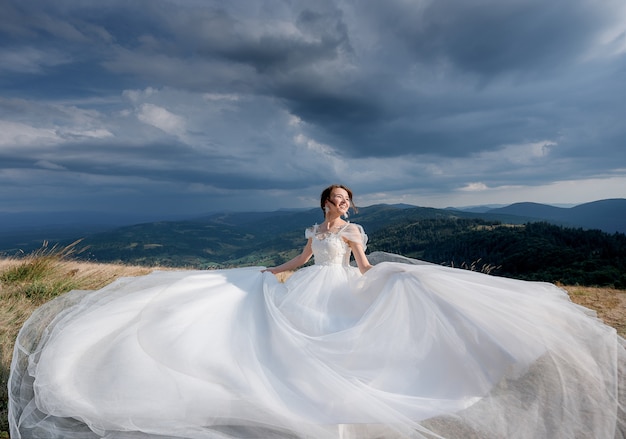 Hermosa novia feliz vestida con un vestido de novia de lujo en el día soleado en las montañas con el cielo nublado