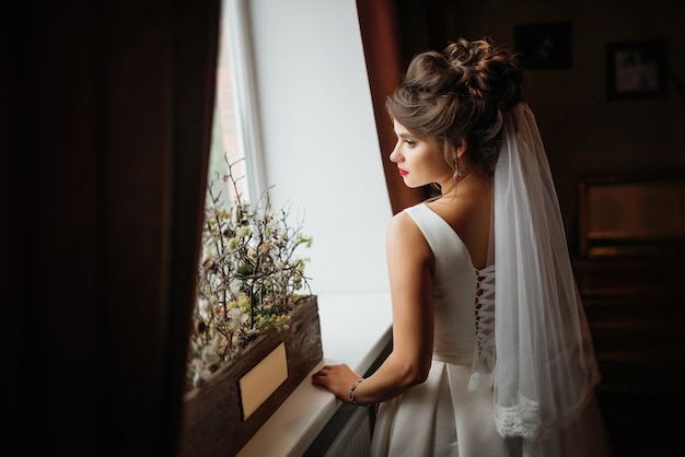 Hermosa novia joven de pie cerca de la ventana en el interior oscuro, su mano en el alféizar de la ventana