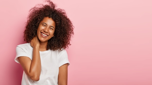 Hermosa novia con mirada divertida tiene una conversación agradable, toca el cuello suavemente, se ríe alegremente de una broma hilarante, está de excelente humor vestida con una camiseta blanca y se encuentra contra una pared rosa con espacio de copia