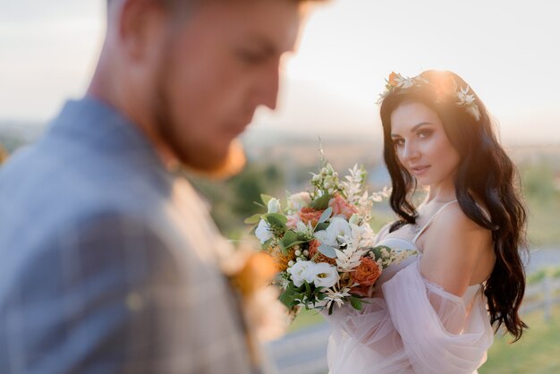 Hermosa novia morena con mirada astuta está sosteniendo un bonito ramo de novia hecho de eustomas frescas y vegetación en la puesta de sol y el novio borroso