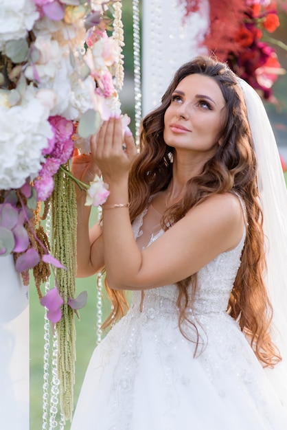 Hermosa novia morena de pelo largo vestida con un vestido de novia cerca del arco de la boda floral
