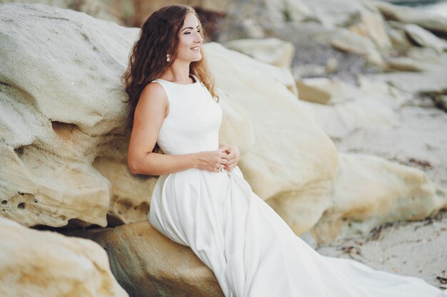 Hermosa novia de pelo largo en un magnífico vestido blanco sentado cerca del agua