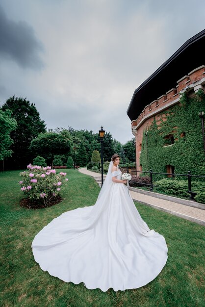 Hermosa novia con ramo de novia y vestido de novia de lujo está de pie delante de un edificio cubierto de hojas verdes