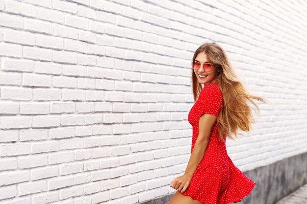 Hermosa novia rubia se vuelve hacia ti y sonriendo, explorando las calles de la ciudad en vacaciones de verano, luciendo feliz, posando junto a la pared de ladrillo blanco al aire libre