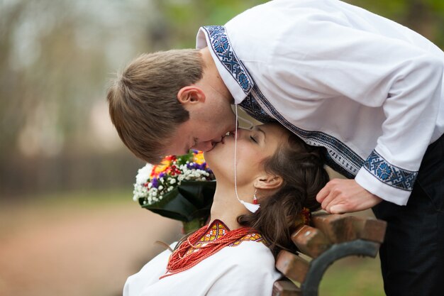 Hermosa novia ucraniana y el novio en trajes de bordado nativo besos en un banco en el fondo de los árboles en un parque