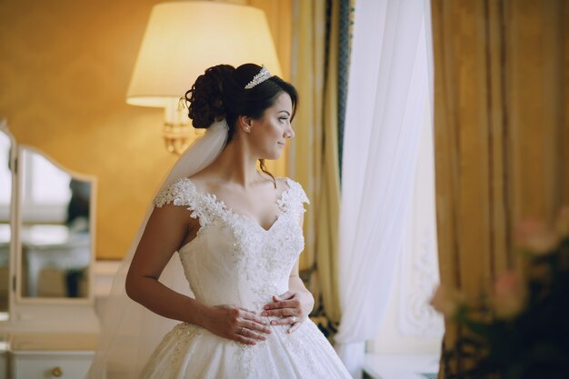 hermosa novia con un vestido blanco y una corona en la cabeza de pie junto a la ventana