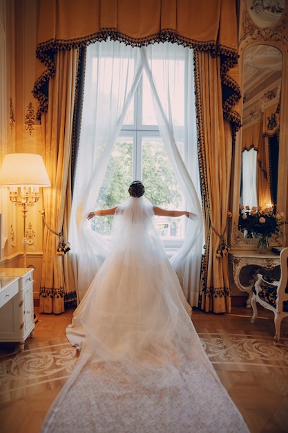 hermosa novia con un vestido blanco y una corona en la cabeza de pie junto a la ventana