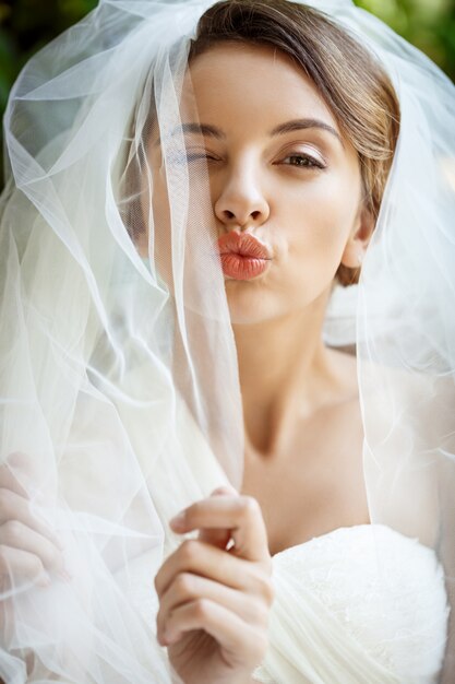 Hermosa novia en vestido de novia y velo guiño, enviando beso.