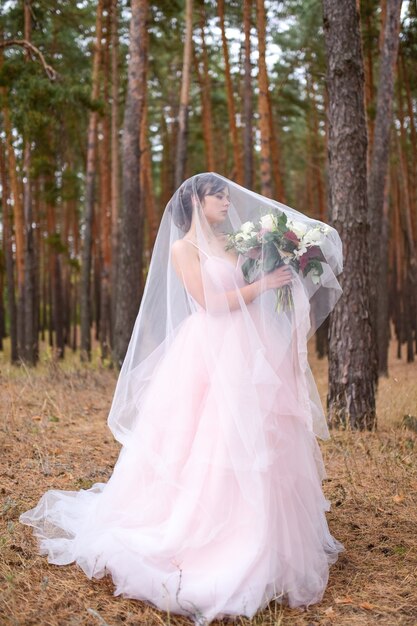 Hermosa novia en vestido rosa está escondida bajo el velo en un bosque