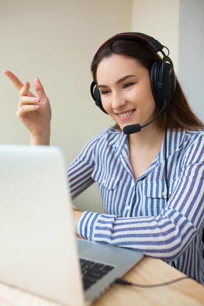 Hermosa operadora independiente que trabaja en marketing en línea con auriculares y una computadora portátil en una computadora de escritorio en la oficina. Chica alegre del centro de llamadas que trabaja desde casa hablando con un cliente