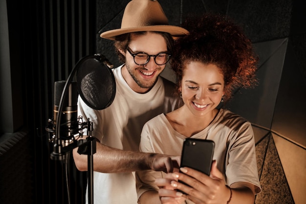 Hermosa pareja de cantantes con estilo felizmente usando el teléfono trabajando juntos en un moderno estudio de grabación de sonido