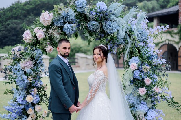 Hermosa pareja caucásica está de pie delante de decorado con arco de hortensias azules y tomados de la mano juntos