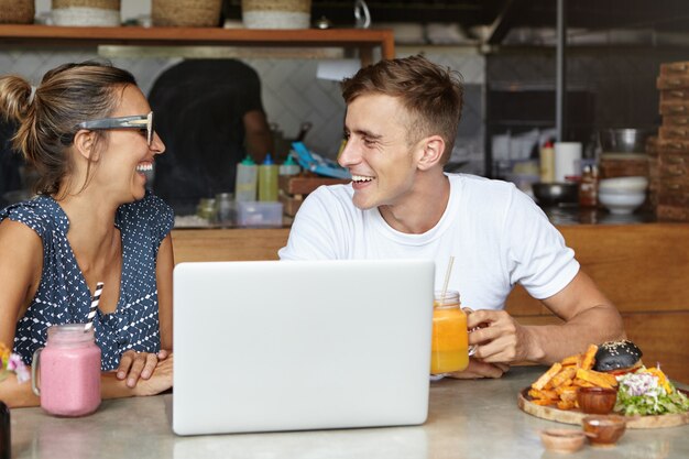 Hermosa pareja conversación animada sentado a la mesa con ordenador portátil y comida en el acogedor interior de la cafetería