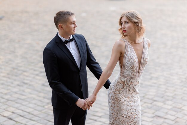 Hermosa pareja de enamorados con elegantes atuendos se mira apasionadamente y se toma de las manos al aire libre