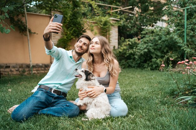 Hermosa pareja feliz haciendo selfie con su adorable perro en el patio trasero mientras está sentado en el césped