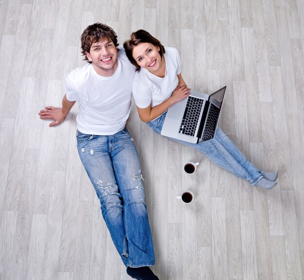 Hermosa pareja feliz sentada en el suelo en casa nueva con portátil