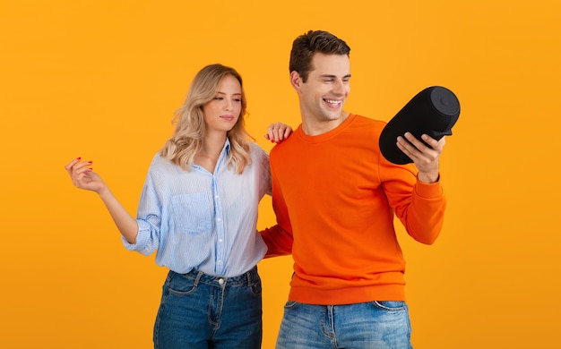 Hermosa pareja joven sonriente sosteniendo altavoz inalámbrico escuchando música bailando en naranja