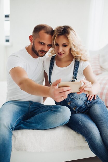 Hermosa pareja de mujeres embarazadas y su esposo vistiendo jeans sentados besándose y tomándose un autorretrato Mamá y papá jóvenes sosteniendo un teléfono inteligente y tomando fotos