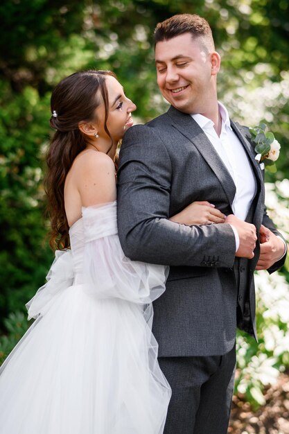 Hermosa pareja de novias novia sonriente en vestido de novia abrazándose detrás de novio guapo mirándose el uno al otro de pie en el jardín Pareja amorosa casada disfrutando del día de la boda