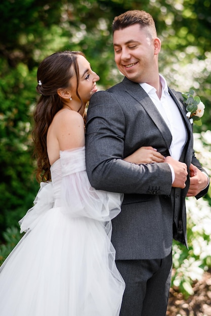 Hermosa pareja de novias novia sonriente en vestido de novia abrazándose detrás de novio guapo mirándose el uno al otro de pie en el jardín Pareja amorosa casada disfrutando del día de la boda