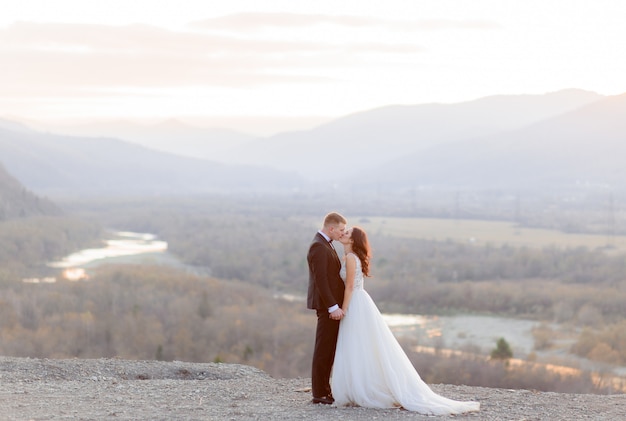 Hermosa pareja de novios se besa en la colina con vistas a un pintoresco paisaje al atardecer