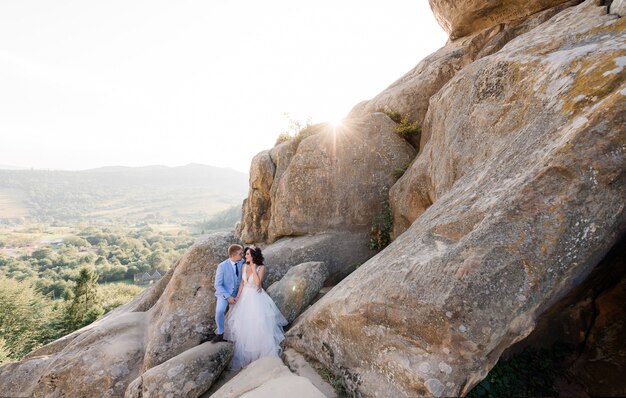 Hermosa pareja de novios en el día soleado está de pie sobre las enormes rocas con vista pintoresca de un bosque