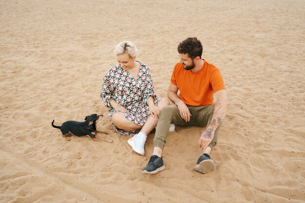 Hermosa pareja relajándose en la arena abrazándose y besándose mientras jugaba con su perro positivo
