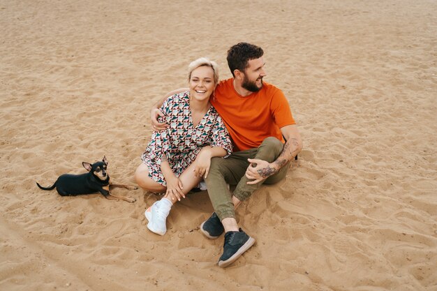 Hermosa pareja relajándose en la arena abrazándose y besándose mientras jugaba con su perro positivo