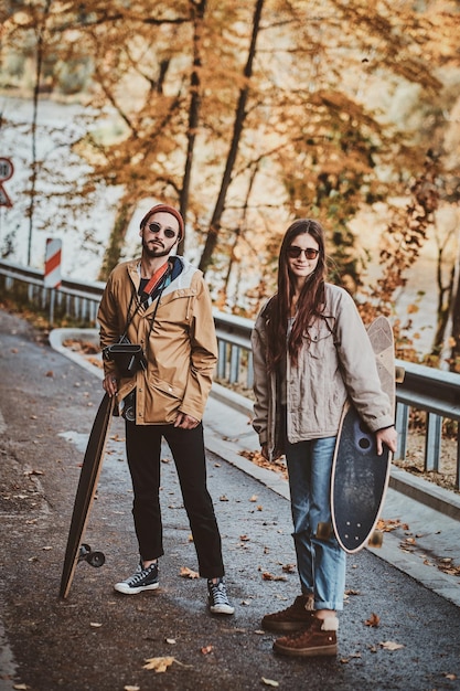 Hermosa pareja romántica con longboards está caminando en el soleado parque de otoño.