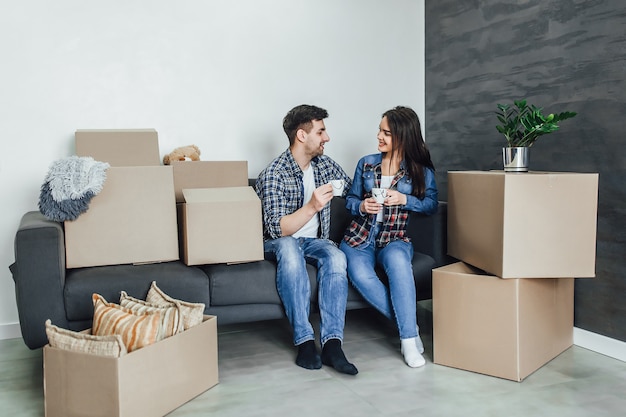 Hermosa pareja en ropa casual está discutiendo el plan de su nueva casa y sonriendo mientras está acostado en el sofá cerca de cajas para mudarse