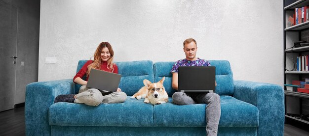 Hermosa pareja sentada en el sofá con Corgi y usando computadoras portátiles