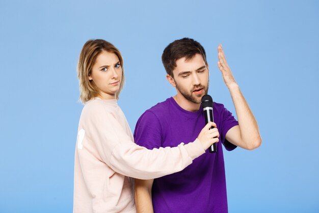 Hermosa pareja sobre pared azul hombre cantando en chica de micrófono disgustado.