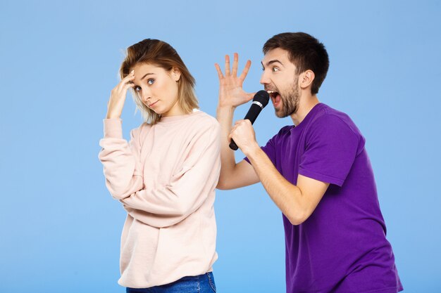 Hermosa pareja sobre pared azul hombre cantando en chica de micrófono disgustado.