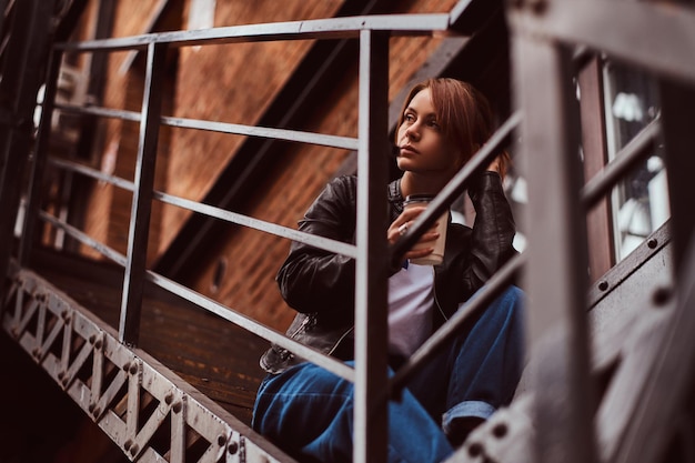 Una hermosa pelirroja con ropa de moda sentada en las escaleras fuera del café y sosteniendo café para llevar.