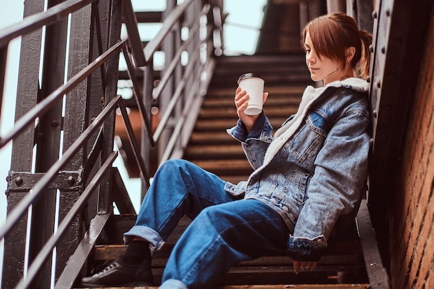 Una hermosa pelirroja con ropa de moda sentada en las escaleras fuera del café y sosteniendo café para llevar.