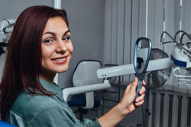 Foto gratuita una hermosa pelirroja sonriente sentada en una silla de dentista con espejo en las manos, mirando la cámara.