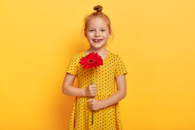 Hermosa pequeña niña de pelo rojo posando con flor en vestido amarillo