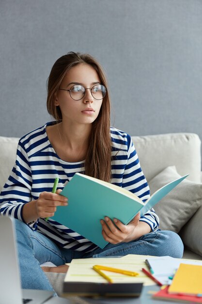 Hermosa periodista reflexiva sostiene un cuaderno y un bolígrafo azul, escribe un artículo, usa gafas redondas transparentes, se sienta en el sofá en casa