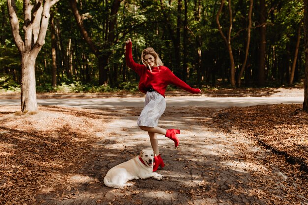 Hermosa rubia feliz en suéter rojo disfrutando de un gran momento con su labrador en el parque. Chica elegante en ropa de abrigo posando entre hojas amarillas caídas.