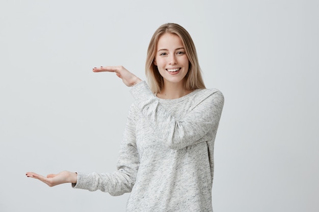 Hermosa rubia guapa con mujer de cabello teñido en ropa casual muestra con las manos la longitud de la caja. Las sonrisas femeninas complacidas demuestran con alegría el tamaño de algo grande. Emociones y sentimientos positivos