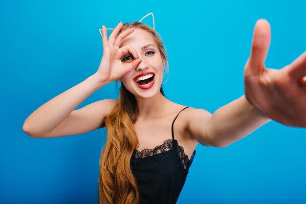 Hermosa rubia con orejas de gato divirtiéndose en la fiesta, sonriendo, tomando selfie. Tiene el pelo largo y ondulado. Con un hermoso vestido negro con encaje, maquillaje brillante.