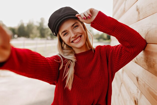 Hermosa rubia en suéter rojo y sombrero negro sonriendo alegremente a su teléfono Hermosa mujer haciendo selfie cerca de la casa de madera en el parque