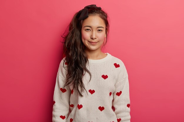 Hermosa tierna chica asiática con cola de caballo larga, mejillas rojas, viste un cómodo jersey con corazones, contra el fondo rosa